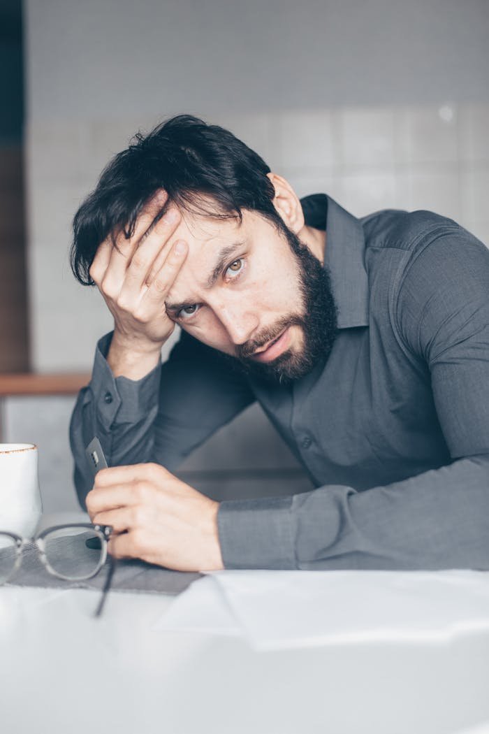 Thoughtful man sitting at a table with glasses and cup, showing frustration.